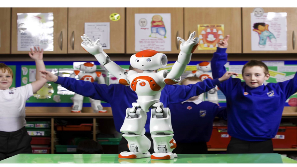 Photo of a small robot on a table with its arms raised, children behind mimicking the pose.