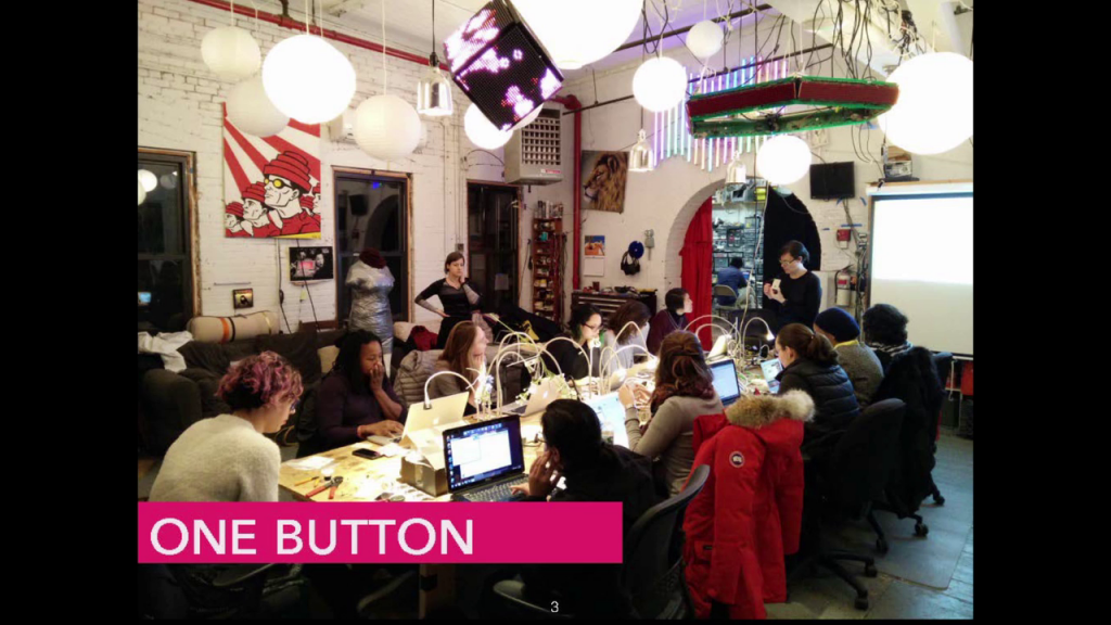 Photo of a dozen women seated around a large table with laptops, as another presents in front of them.