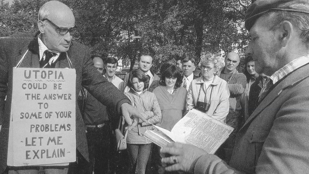 An older man with a placard haging from his neck reading "Utopia could be the answer to some of your problems. Let me explain." surrounded by a crowd of onlookers.