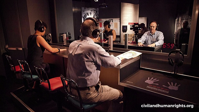 A man and woman sitting at a simulated lunch counter facing a mirror. They are wearing headphones and have their hands placed flat on the counter