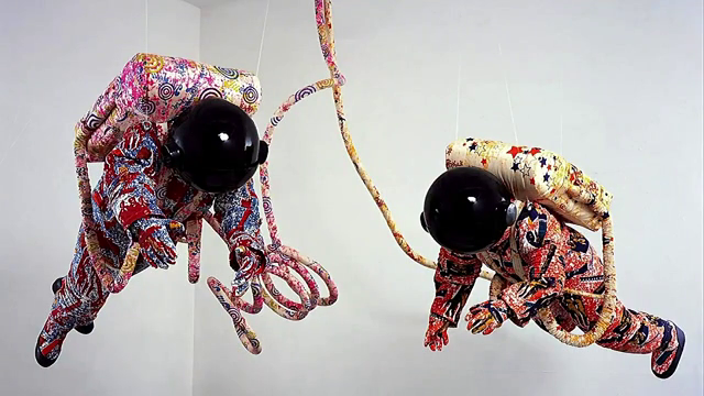 Approximations of astronaut suits made of heavily-patterned fabric hanging from the ceiling of a gallery space.