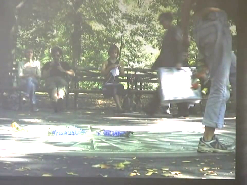 Kristen knitting while seated on a park bench, various people nearby or walking by