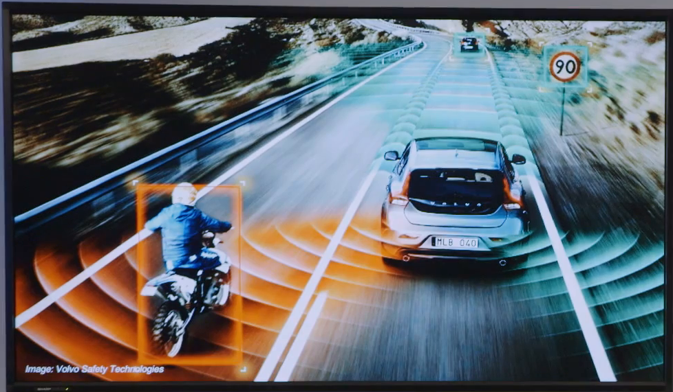 Video still showing a car on a highway with a motorcycle following closely behind. Waves are illustrated emanating from the car,, red where they meet the motorcycle rider, indicating a detection of proximity