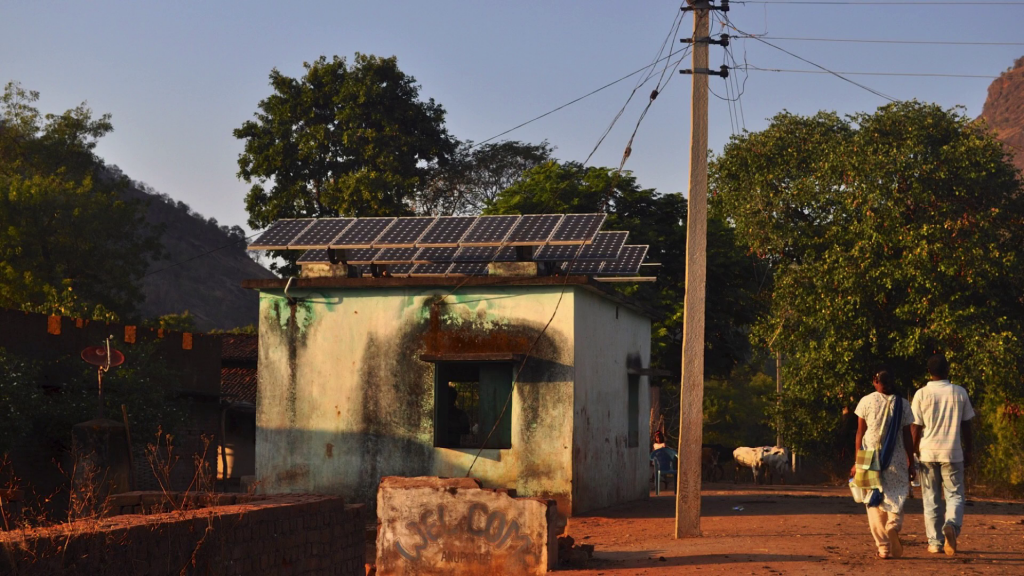 Two people walking in a rural area near a small building with solar panels on its roof