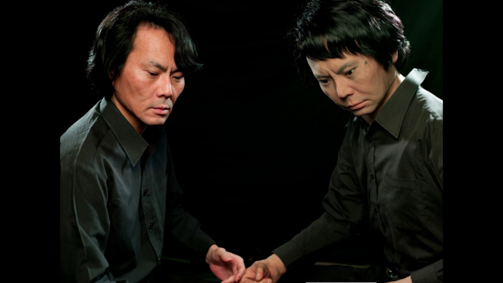 Hiroshi Ishiguro, shown from the waist up leaning slightly forward with his hand in front of him, mirrored by a robot modeled on his features