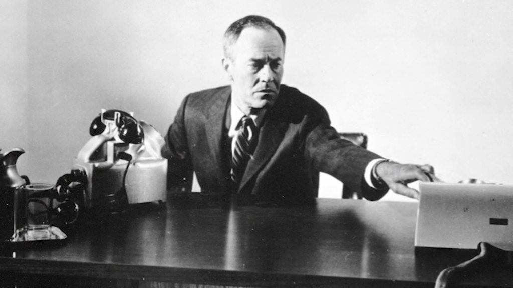 A man seated at an office desk, reaching to push a button on a machine on the desktop