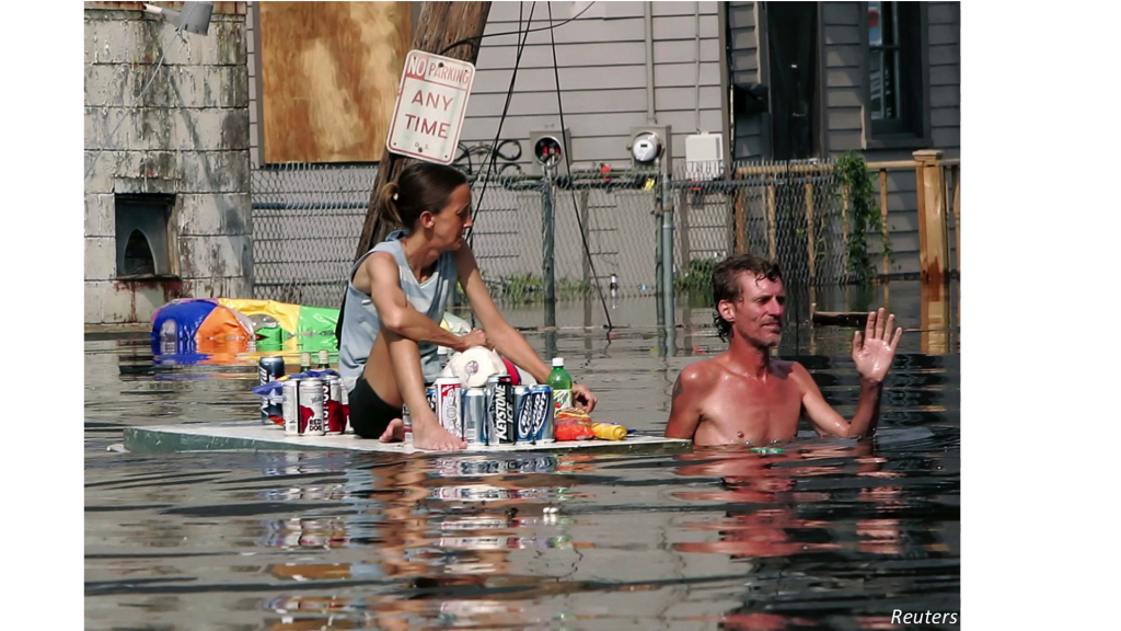 A man in chest-deep water pulling along a makeshift raft with a woman on it surrounded by some supplies, though mostly cans of beer.