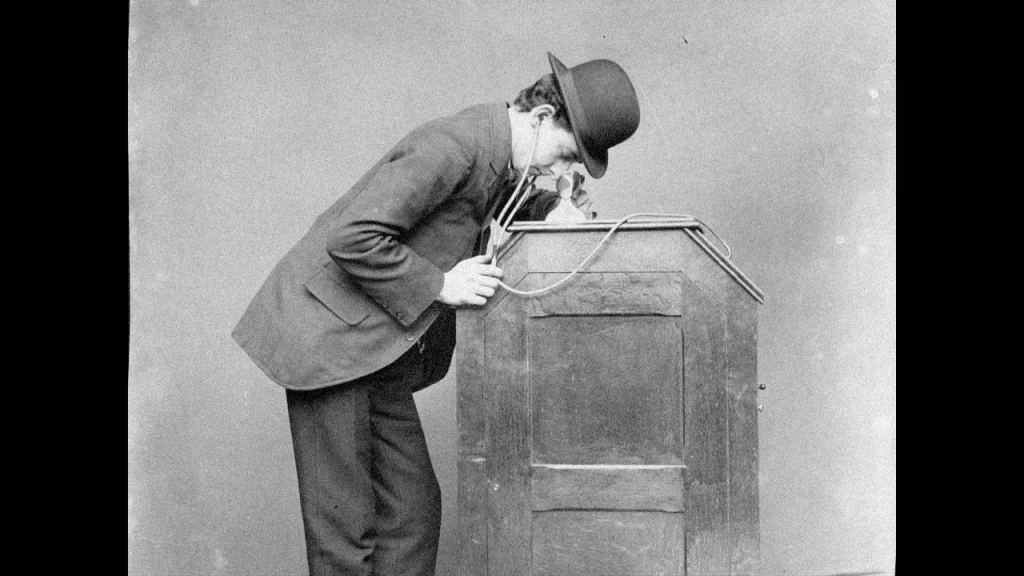 A man leaning over a wooden cabinet, peering into the top of it, with earpieces for hearing audio in his ears.
