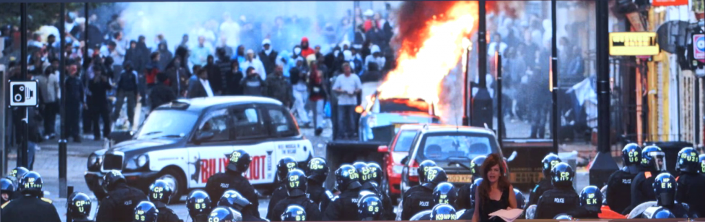 Photo of a group of policemen in riot gear approaching a large crowd of rioters, a burning car in the space between them