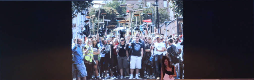 A group of people standing in the middle of a street, holding brooms bristles-up above their heads