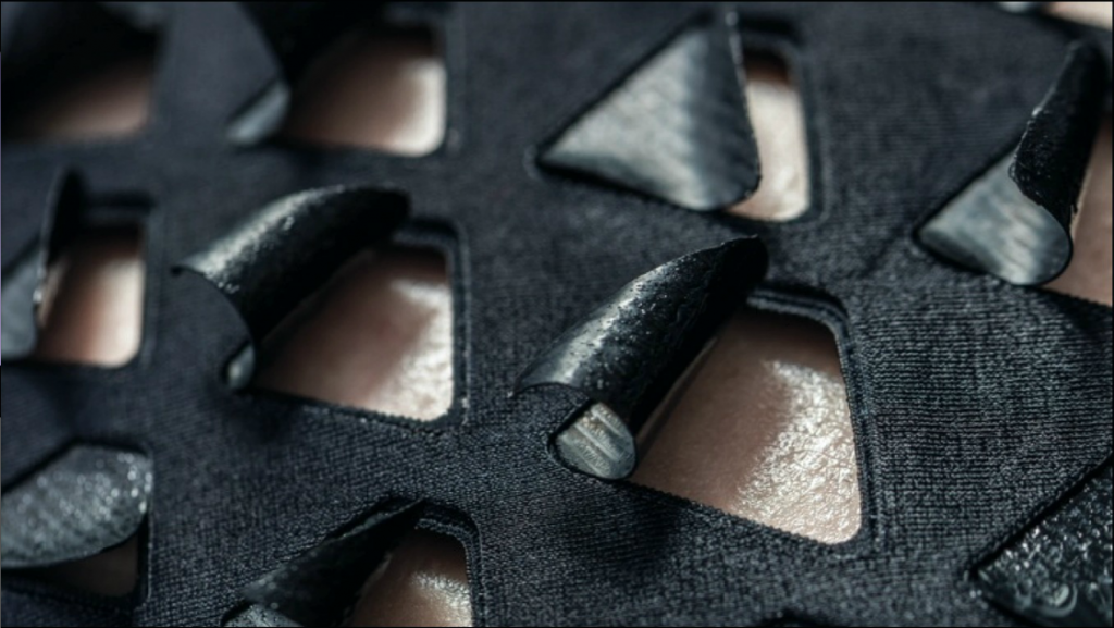 Close-up of a fabric with trapezoidal openings in it with flaps of a matching shape curled away from them, skin visible below