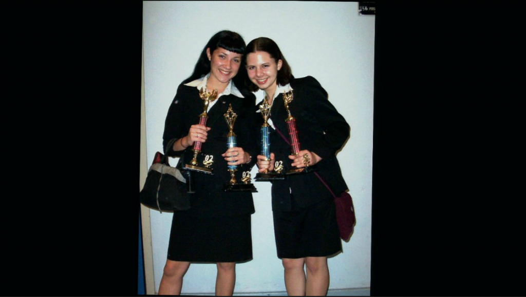 Two teenage girls in school uniforms standing with their heads close together, holding several trophies