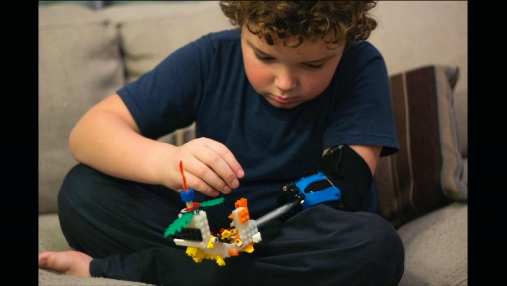 Photo of a young boy with a prosthetic arm attachment, placing Lego pieces on a custom end piece.