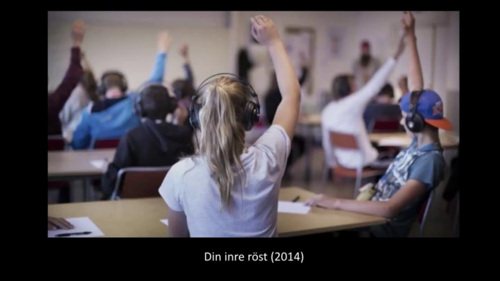 A classroom full of children wearing headphones, all holding their hands up in the air