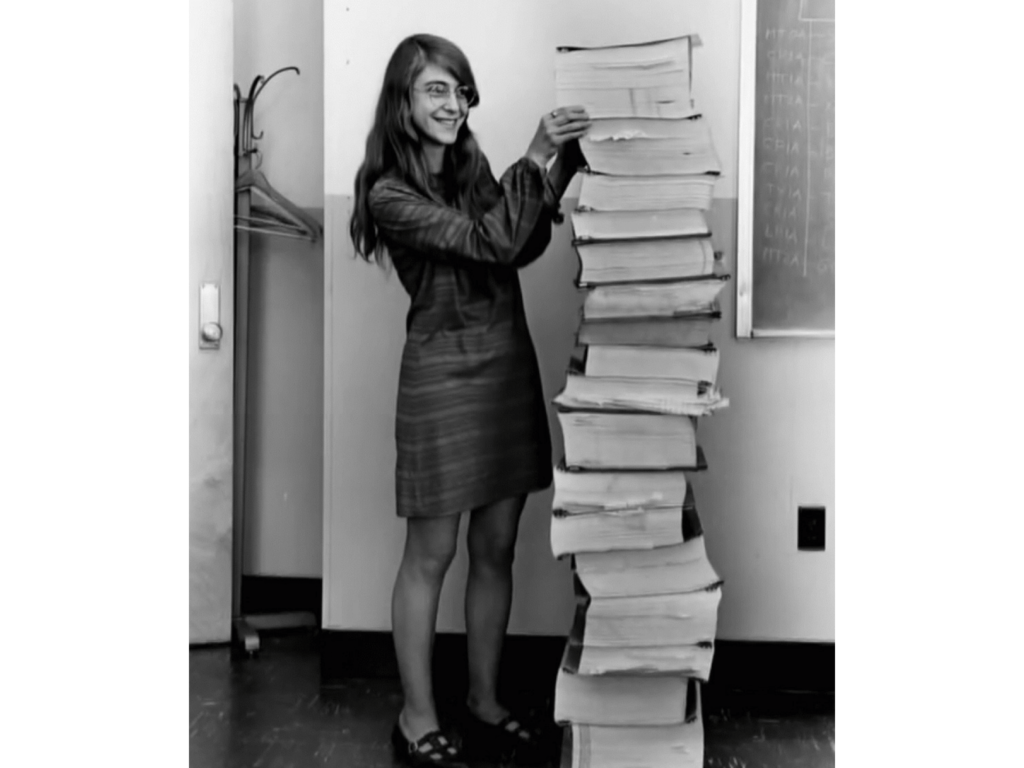 A young Margaret Hamilton, smiling, propping up a stack of paper as tall as herself