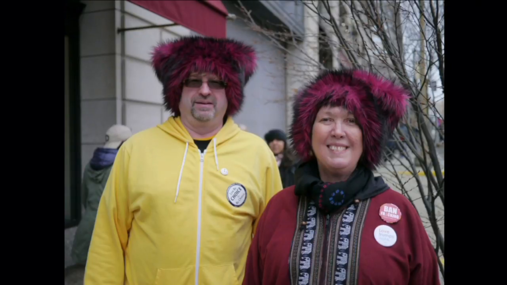 A man and woman wearing a variation of the pussyhat made of dark red and black fake fur