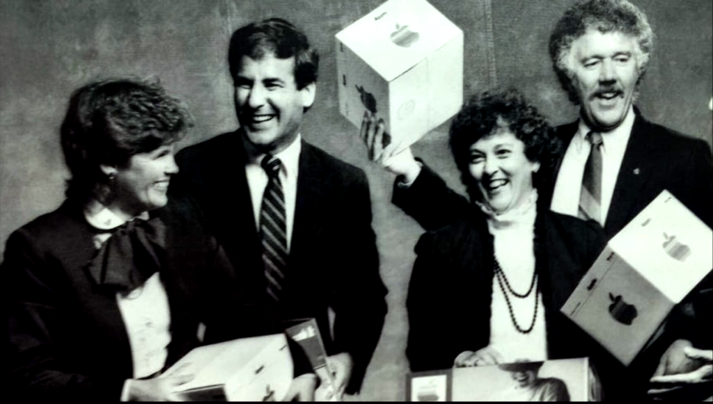 Two men and two women in office dress smiling and holding Apple product boxes