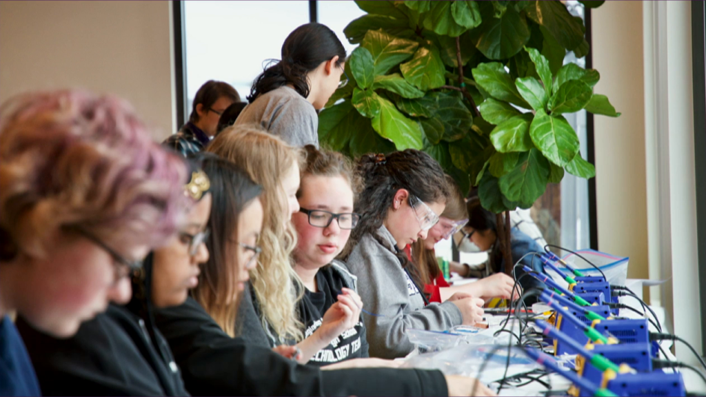 A table full of young women, some wearing safety glasses, seated before electronics testing equipment.