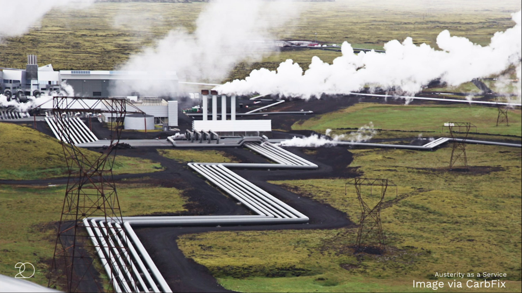 A wide landscape with pipes zig-zagging across the terrain leading toward a large factory-style building billowing large clouds of white vapor