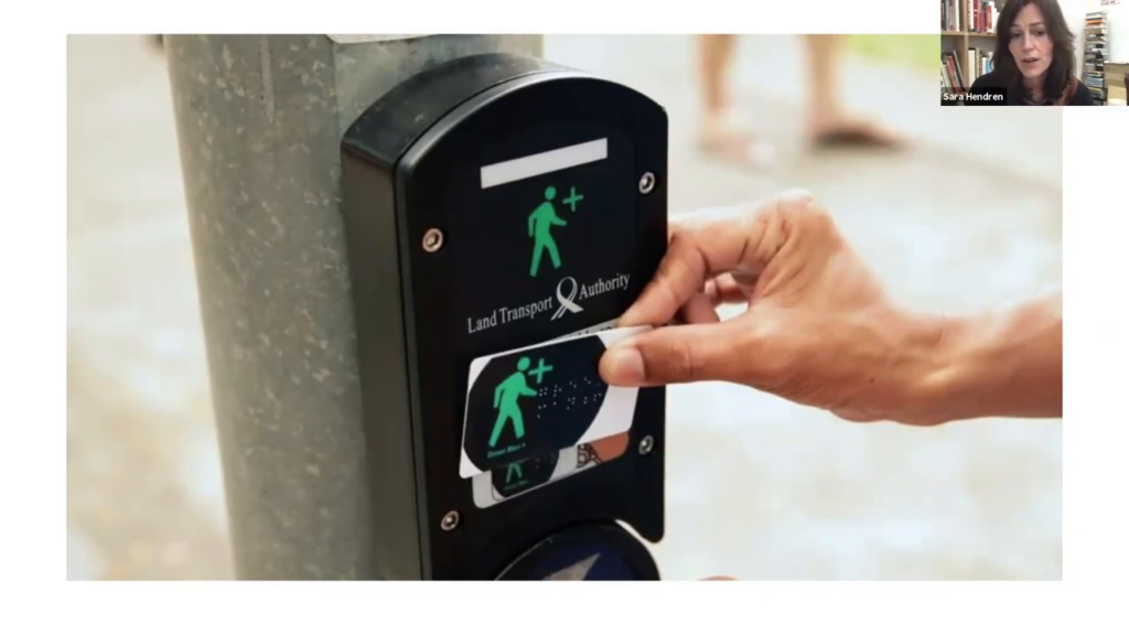 A crossing signal request box with an illustration of a green-colored walking figure, and a hand touching a card with the same symbol to the box.